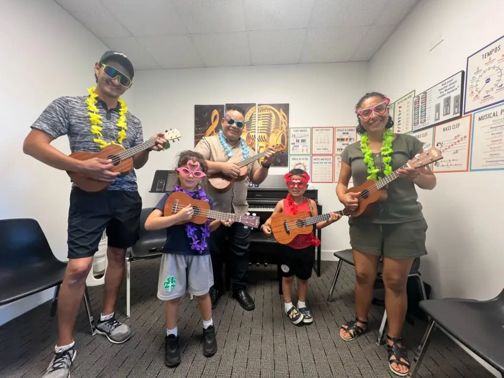 A group of five people, including two children and three adults, stand in a music room at Music Lessons Westchase wearing colorful leis and novelty glasses, each holding a ukulele and smiling at the camera. Music posters and a piano are in the background.