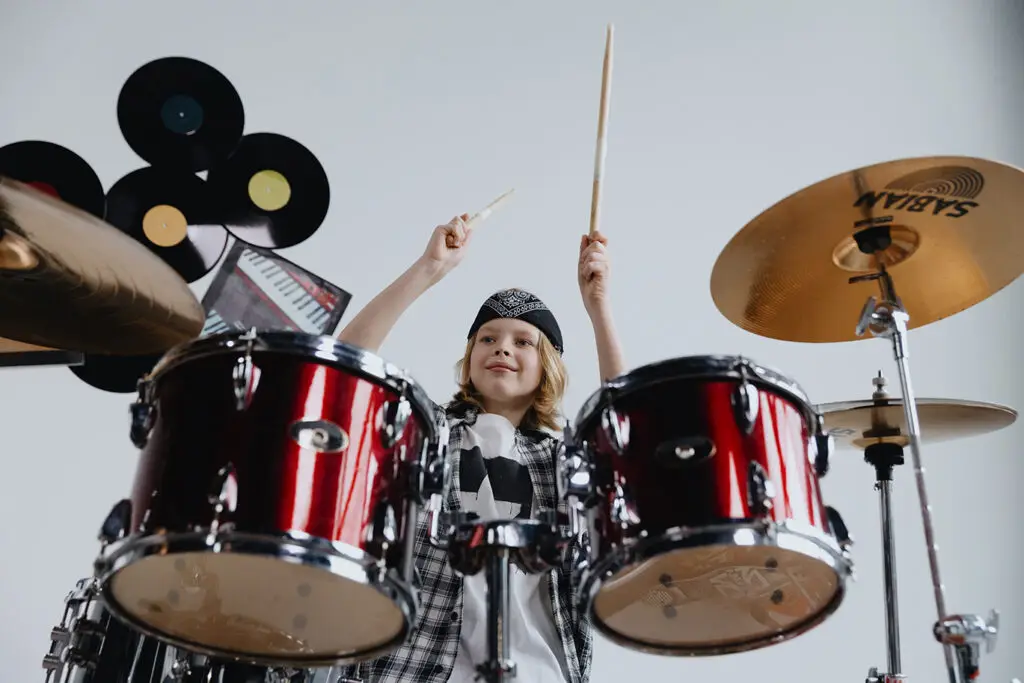 A child wearing a bandana plays a red drum set enthusiastically, holding drumsticks in the air. Vinyl records and a keyboard are mounted on a white wall in the background—perfect inspiration for music lessons Westchase families will love.