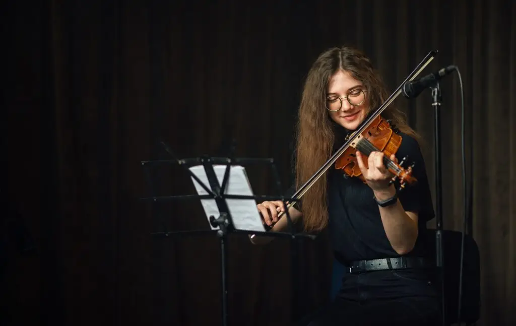A young woman with long brown hair and glasses plays the violin while reading sheet music on a stand, sitting in front of a microphone on a dimly lit stage—showcasing skills honed from music lessons Westchase offers.