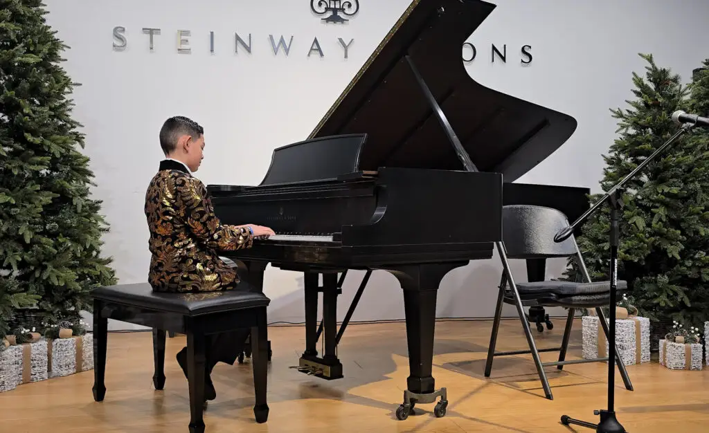 A young boy in a gold and black jacket plays a grand piano on stage, surrounded by decorated pine trees—an inspiring scene from music lessons Westchase. A microphone stand and empty chair are nearby, with STEINWAY visible on the wall behind him.