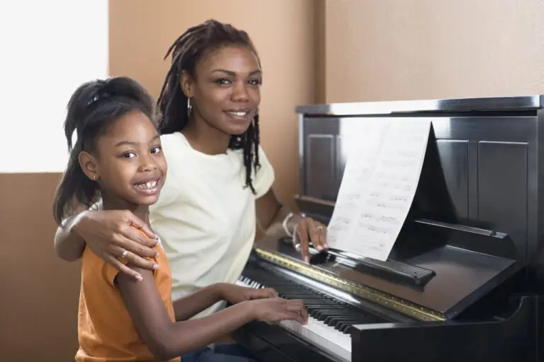 A woman and a young girl sit together at a piano during music lessons Westchase. The woman smiles while holding the girl, who is playing the keys. Sheet music rests on the piano, and both look happily at the camera.