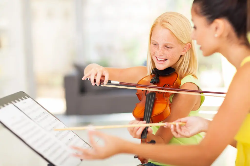 A smiling young girl plays the violin while her teacher points to sheet music on a stand in a bright, indoor setting—capturing the joy of music lessons Westchase families love.