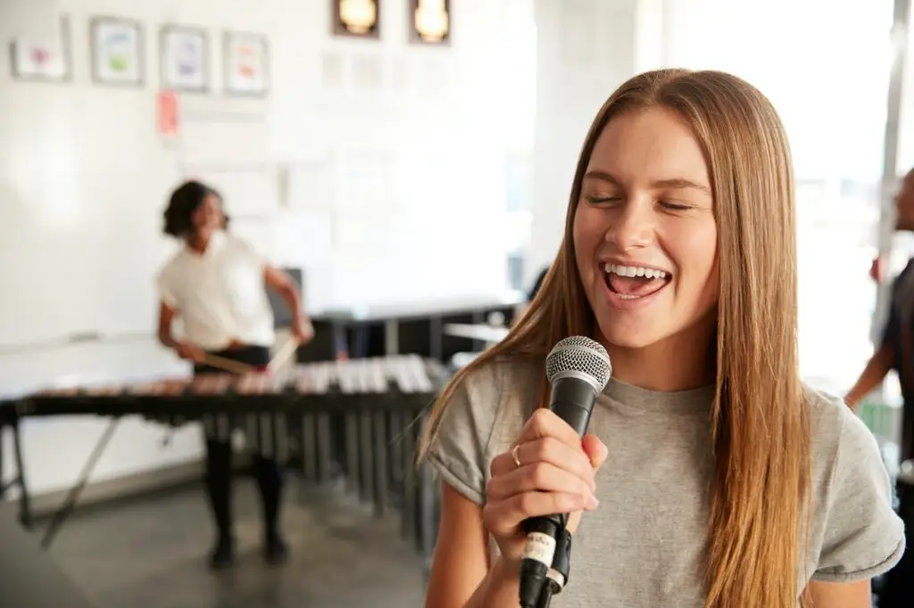 A young woman with long hair sings into a microphone, eyes closed and smiling, during music lessons in Westchase, while another person plays a xylophone in the blurred background of a bright classroom.