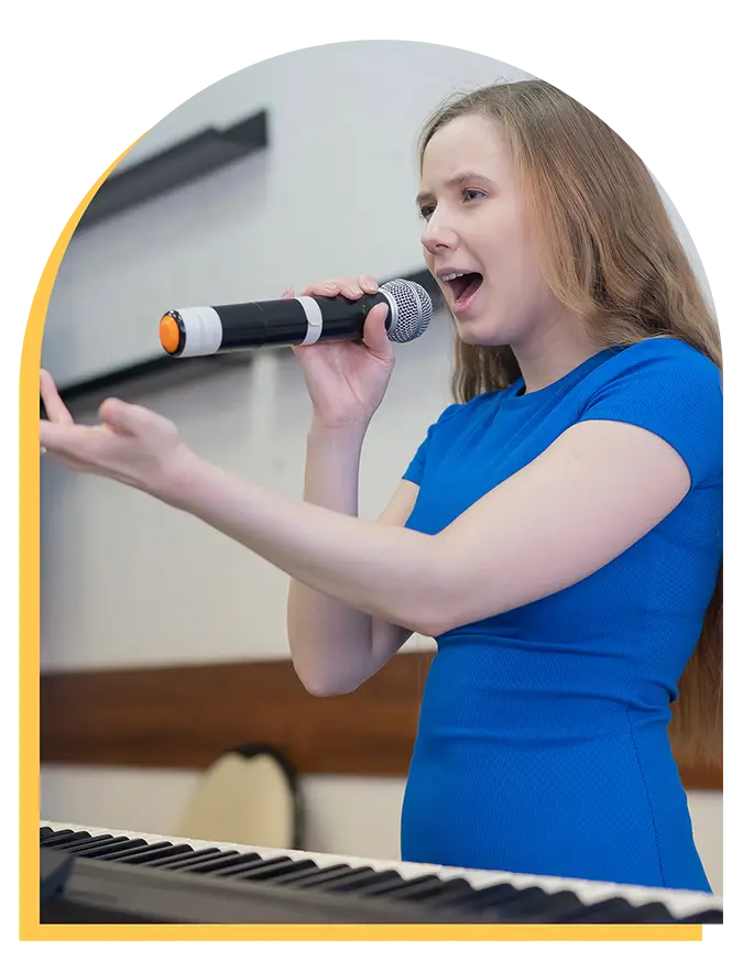 A woman in a blue dress sings passionately into a microphone while standing behind a keyboard, with one hand gesturing expressively, showcasing the talent you can develop through music lessons Westchase offers.
