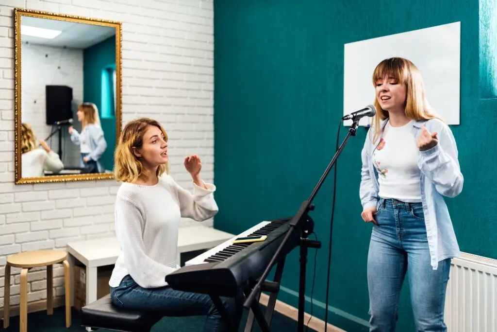 A woman plays a keyboard while another woman sings into a microphone in a music studio with teal walls and a large mirror, capturing the creative spirit of music lessons Westchase.
