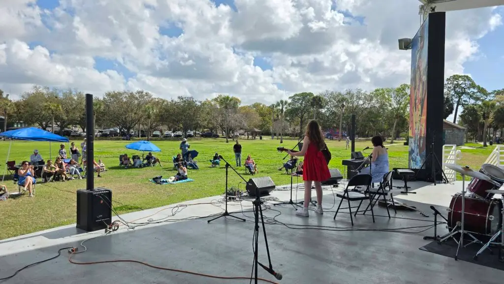 A band performs on an outdoor stage facing a grassy field, with a girl in a red dress playing guitar—showcasing skills that could be learned through music lessons Westchase. Audience members relax on blankets under a partly cloudy sky.