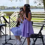 A young girl in a purple dress sings into a microphone on an outdoor stage, while another person in a striped dress plays guitar beside her; trees and water are visible in the background.