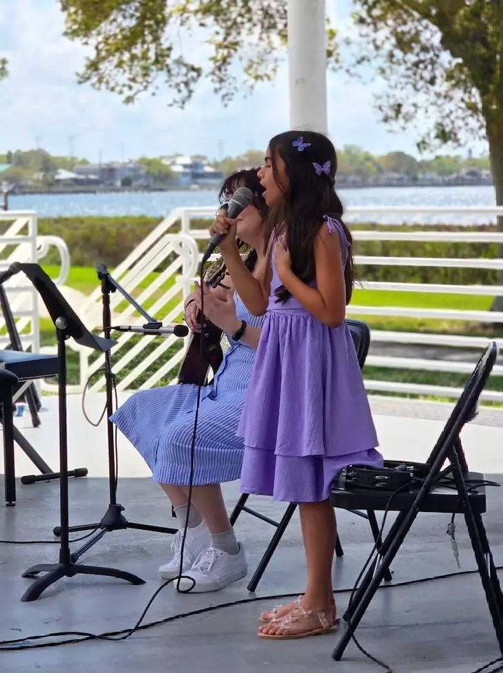 A young girl in a purple dress sings into a microphone on an outdoor stage, while another person in a striped dress plays guitar beside her; trees and water are visible in the background.