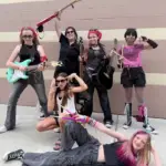 Six young women dressed in rock-inspired outfits pose with electric guitars and drumsticks in front of a tan brick wall. Two sit on the ground, while four stand behind them, all looking energetic and playful.