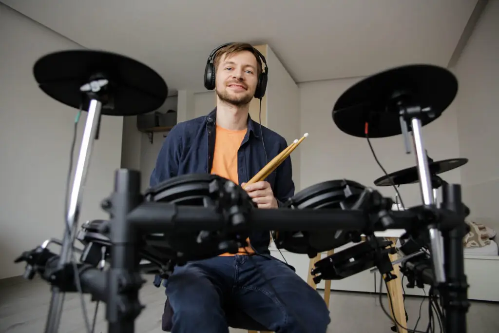 Portrait of smiling young man with headphones hitting electric drum set during repetition at home. Online music lessons.