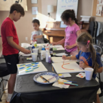Four children are gathered around a table covered with art supplies, painting on canvases in a brightly lit room decorated with musical notes and an ART CLASS sign on the wall.