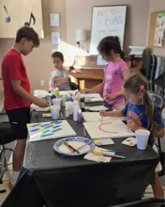 Four children are gathered around a table covered with art supplies, painting on canvases in a brightly lit room decorated with musical notes and an ART CLASS sign on the wall.