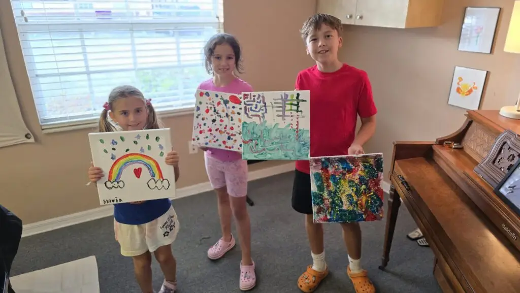 Three children stand indoors, each holding a colorful painting. The left child’s painting features a rainbow, the middle child’s has colorful dots and lines, and the right child’s canvas shows abstract patterns. They are smiling.