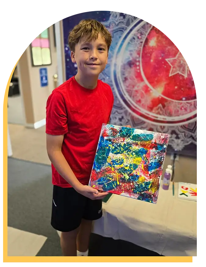 A smiling boy in a red shirt and black shorts holds a colorful abstract painting. He stands indoors near a table and in front of a large, decorative wall tapestry with intricate patterns and a red star design.