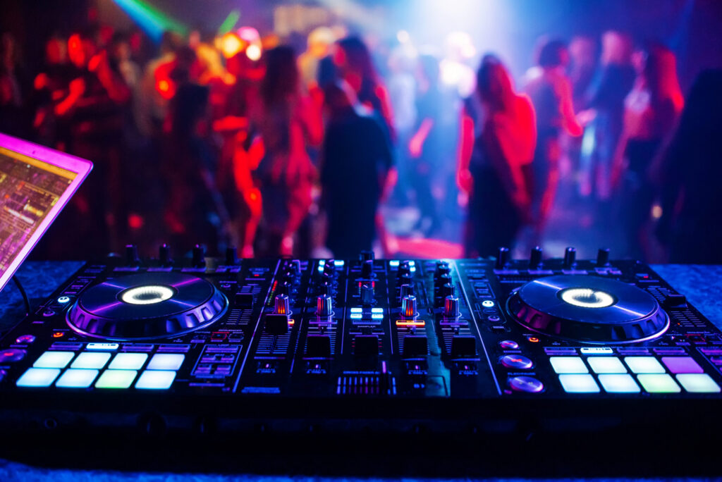 A close-up of a DJ mixing console with glowing buttons and dials, set on a table in a nightclub. In the background, colorful lights illuminate a crowd of people dancing.
