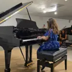 A woman in a blue dress plays a grand piano on a stage while an audience watches from the background. The setting appears to be an indoor recital or music event.