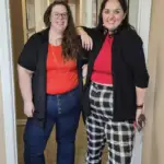 Two women smiling and standing in a hallway. Both wear red tops and black outer layers; one has blue jeans and glasses, the other has black-and-white plaid pants and holds a face mask.