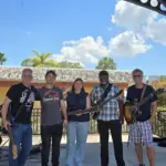 Five people stand on an outdoor stage with guitars and a bass, smiling at the camera under a sunny sky with clouds. Trees and a building are visible in the background.