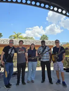 Five people stand on an outdoor stage with guitars and a bass, smiling at the camera under a sunny sky with clouds. Trees and a building are visible in the background.
