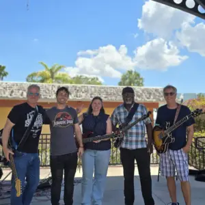 Five people stand on an outdoor stage with guitars and a bass, smiling at the camera under a sunny sky with clouds. Trees and a building are visible in the background.